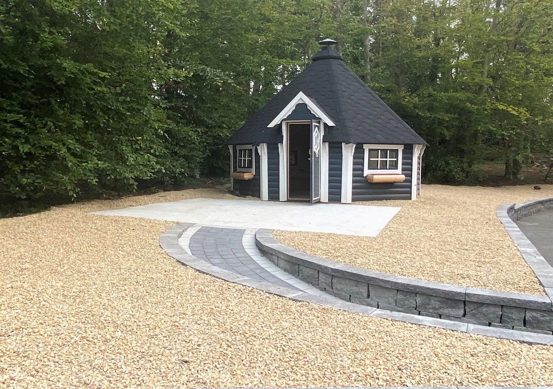 Hexagonal dark timber BBQ hut with white trim surrounded by gravel and trees | Signature Garden Rooms, Central Scotland